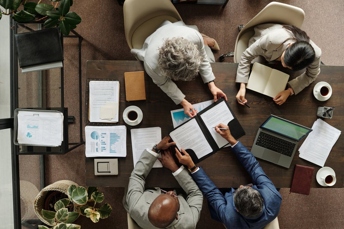 Group of middle aged multiethnic business professionals collaborating around table, reviewing documents and using laptop