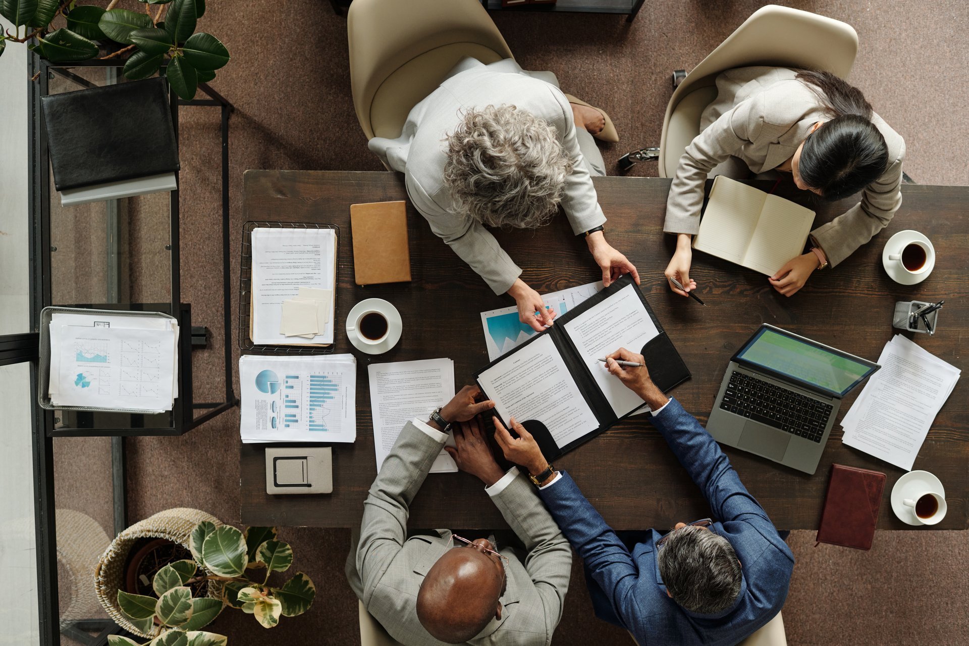 Group of middle aged multiethnic business professionals collaborating around table, reviewing documents and using laptop
